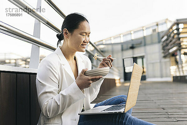Glückliche Geschäftsfrau  die Salat isst und mit einem Laptop sitzt