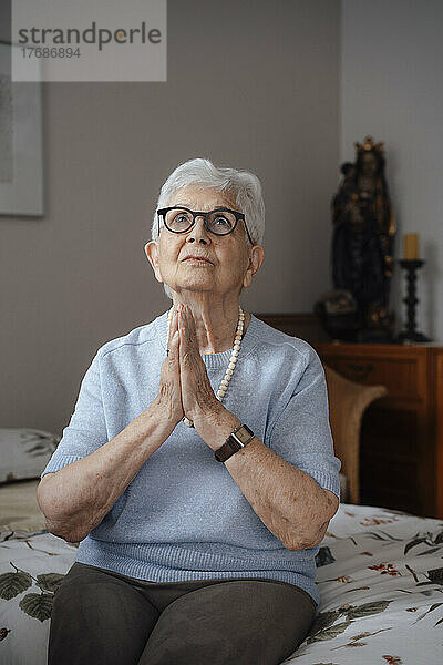 Ältere Frau mit gefalteten Händen sitzt zu Hause auf dem Bett
