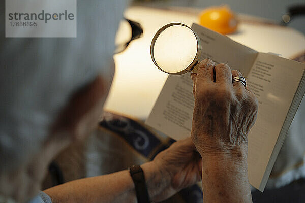 Ältere Frau mit Lupe liest Buch zu Hause