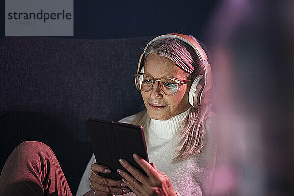 Ältere Frau mit Kopfhörern und Tablet-PC sitzt auf einem Stuhl