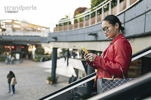 Junge Frau mit Einkaufstasche benutzt Smartphone und fährt auf Rolltreppe nach unten