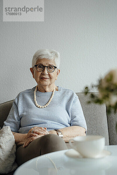 Ältere Frau mit gefalteten Händen sitzt zu Hause auf dem Sofa