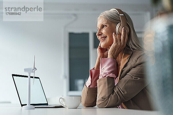 Lächelnde Geschäftsfrau hört Musik über Kopfhörer und sitzt am Schreibtisch im Büro