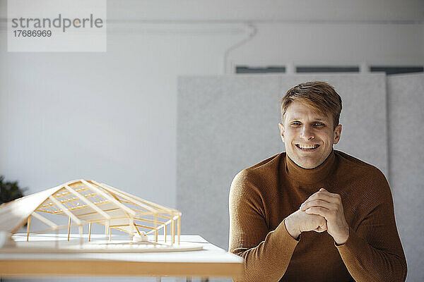 Glücklicher Architekt mit gefalteten Händen  der neben einem blattförmigen Holzmodell im Büro sitzt