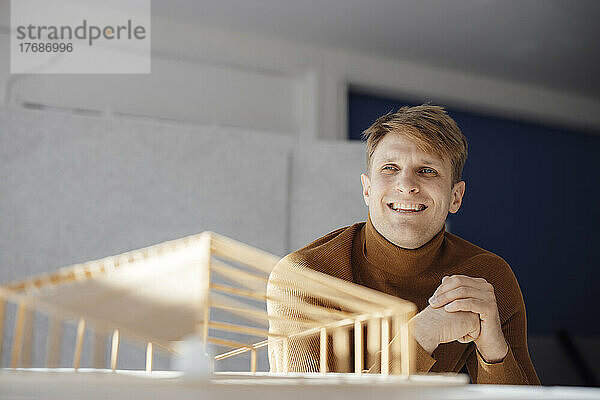 Lächelnder Architekt mit gefalteten Händen sitzt neben einem blattförmigen Holzmodell im Büro