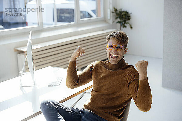Happy businessman wearing eyeglasses gesturing fist sitting at desk in office