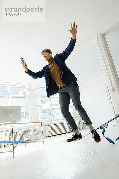 Lächelnder Geschäftsmann  der auf dem Seil balanciert und im Büro ein Selfie mit dem Smartphone macht