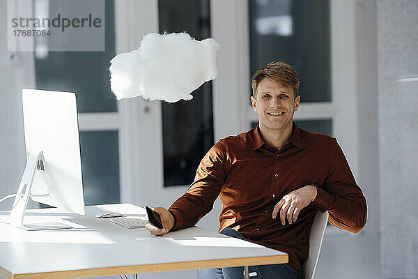 Glücklicher Geschäftsmann mit Mobiltelefon und schwebendem Cloud-Netzwerk im Büro