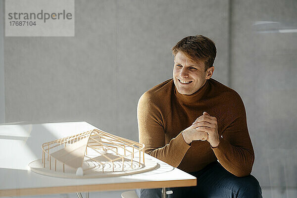 Glücklicher Architekt sitzt mit blattförmigem Modell am Schreibtisch im Büro