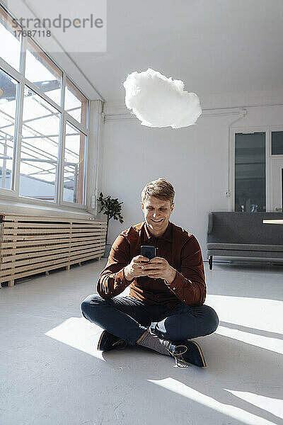 Lächelnder Geschäftsmann  der im Büro unter einem schwebenden Cloud-Netzwerk sitzt und sein Smartphone benutzt