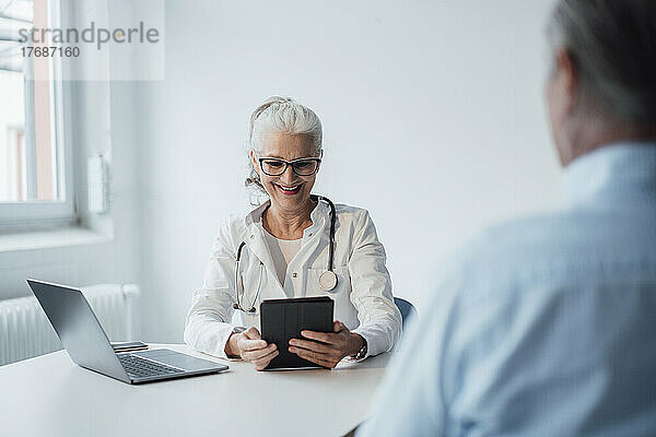 Glücklicher Arzt  der einen Tablet-PC verwendet und mit dem Patienten am Schreibtisch sitzt