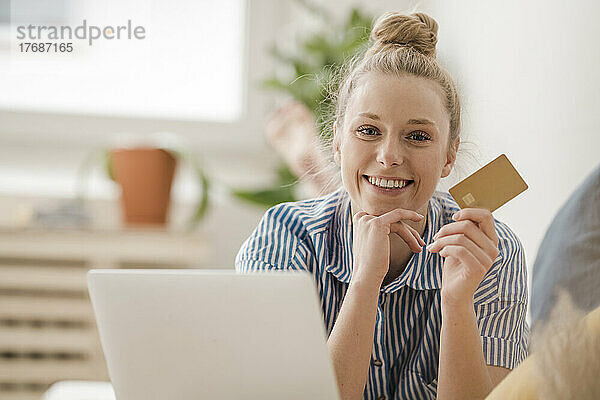 Lächelnde Geschäftsfrau mit der Hand am Kinn  die zu Hause ihre Kreditkarte zeigt