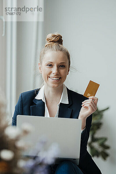 Lächelnde Geschäftsfrau mit Laptop zeigt Kreditkarte im Büro