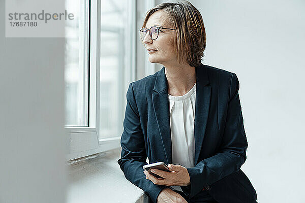Smiling businesswoman with smart phone looking out through window