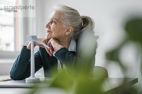 Nachdenkliche Geschäftsfrau mit Windturbinenmodell sitzt am Schreibtisch im Büro