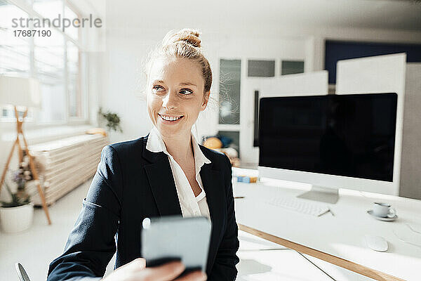 Lächelnde Geschäftsfrau  die darüber nachdenkt  ihr Smartphone im Büro zu halten