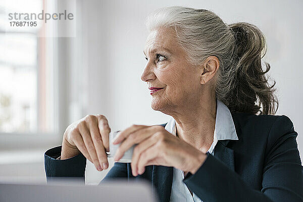 Lächelnde Geschäftsfrau mit Kaffeetasse sitzt im Büro