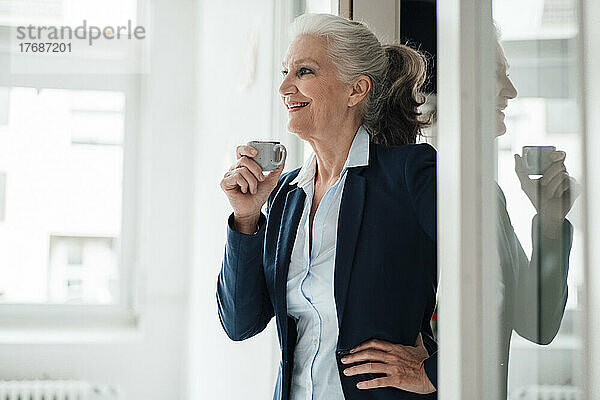 Glückliche Geschäftsfrau hält Kaffeetasse und steht mit der Hand auf der Hüfte im Büro