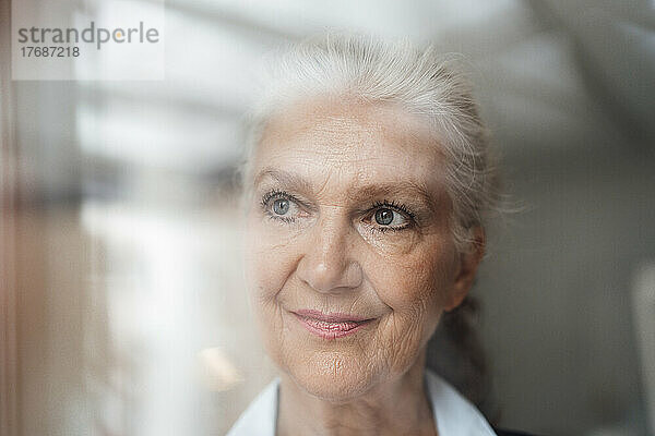 Lächelnde ältere Geschäftsfrau mit grauem Haar  die durch das Fenster schaut