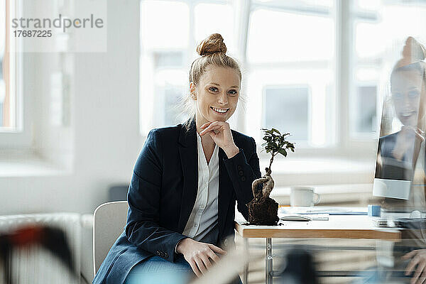Lächelnde Geschäftsfrau sitzt mit Pflanze auf dem Schreibtisch im Büro