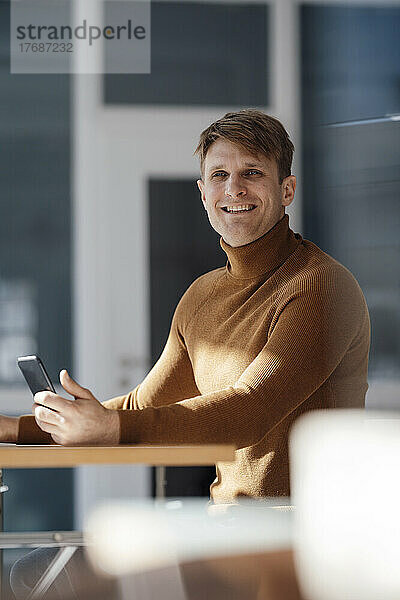Glücklicher Geschäftsmann mit Mobiltelefon am Schreibtisch im Büro