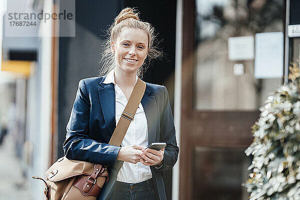 Lächelnde Geschäftsfrau mit Umhängetasche und Smartphone