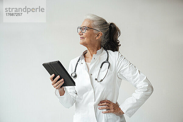 Arzt hält Tablet-PC stehend mit der Hand auf der Hüfte vor weißem Hintergrund