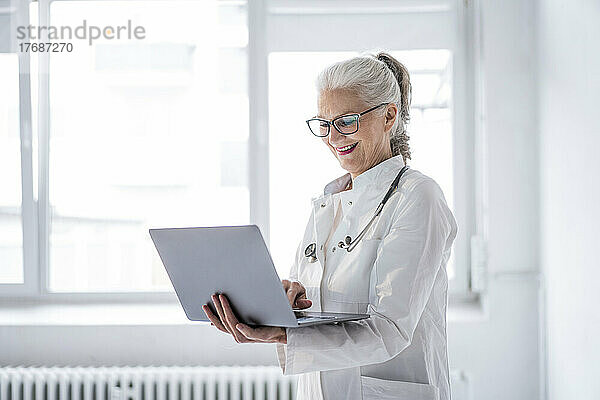Glücklicher Oberarzt mit Laptop  der am Fenster steht