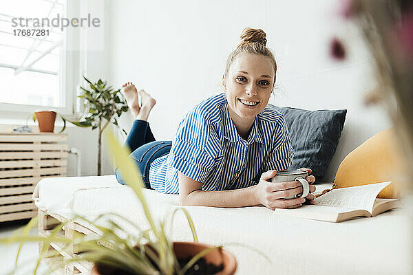 Lächelnder Freiberufler mit Kaffeetasse und Buch zu Hause im Bett liegend