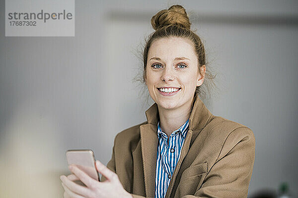 Lächelnde Geschäftsfrau im Blazer mit Smartphone im Büro