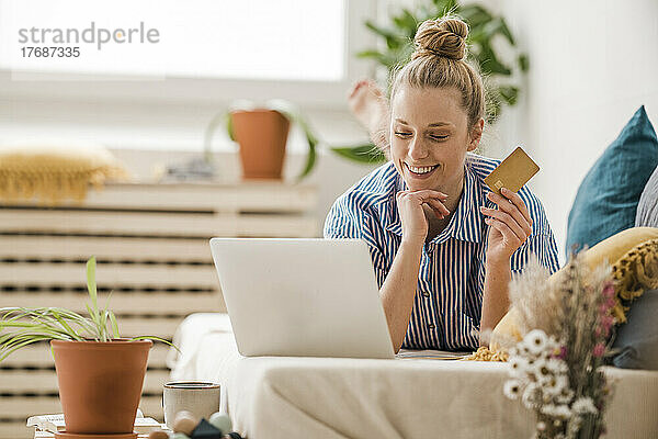 Lächelnder Freiberufler mit Kreditkarte und Blick auf den Laptop  der zu Hause im Bett liegt
