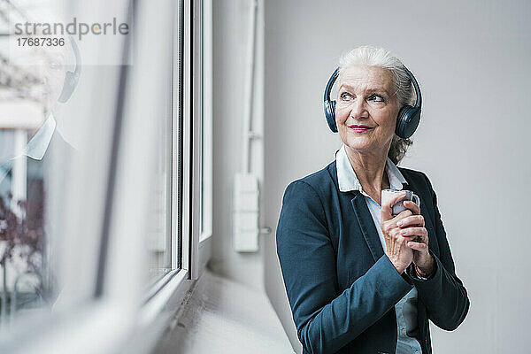 Geschäftsfrau mit kabellosen Kopfhörern steht am Fenster im Büro