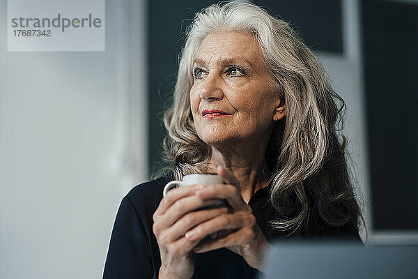 Nachdenkliche ältere Geschäftsfrau mit Kaffeetasse sitzt im Büro