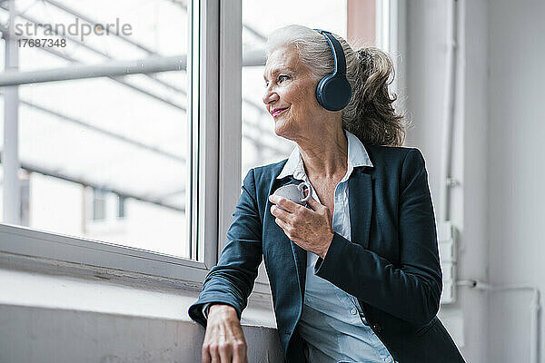 Lächelnde Geschäftsfrau hört Musik über kabellose Kopfhörer  die am Fenster im Büro steht