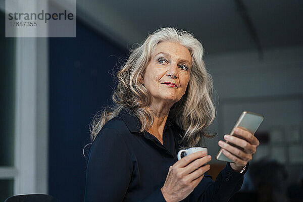 Ältere Geschäftsfrau hält Kaffeetasse und Smartphone im Büro