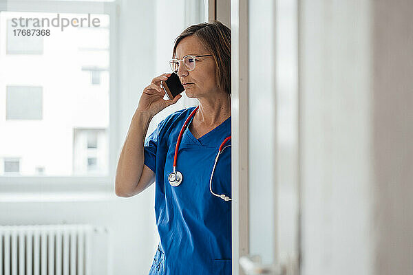 Arzt spricht mit Smartphone und steht im Türrahmen der Klinik
