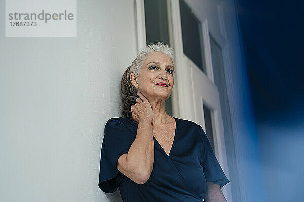 Lächelnde ältere Frau mit grauen Haaren steht an der Wand im Büro