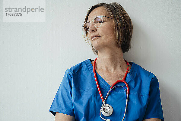 Erschöpfte Ärztin mit geschlossenen Augen lehnt an einer weißen Wand und ruht im Krankenhaus