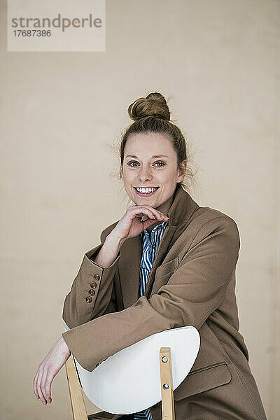 Lächelnde Geschäftsfrau mit der Hand am Kinn sitzt auf einem Stuhl vor der Wand