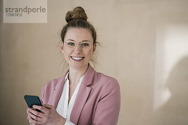 Glückliche junge Geschäftsfrau hält Smartphone vor der Wand