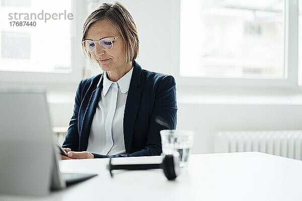 Lächelnde Geschäftsfrau  die am Schreibtisch im Büro sitzt und ihr Mobiltelefon benutzt