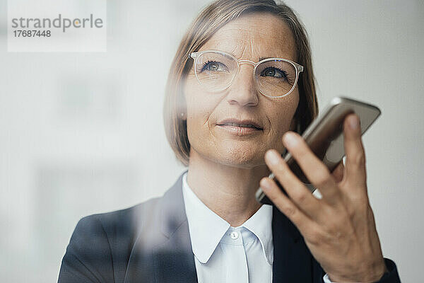 Reife Geschäftsfrau mit Brille spricht über den Lautsprecher ihres Mobiltelefons