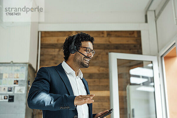 Glücklicher Geschäftsmann  der im Büro über ein kabelloses Headset auf dem Smartphone spricht