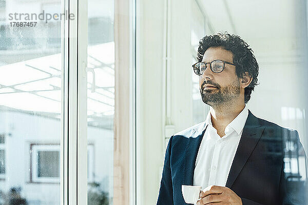 Nachdenklicher Geschäftsmann mit Kaffeetasse und Blick durch das Fenster im Büro