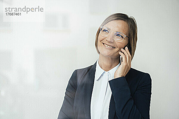 Glückliche Geschäftsfrau im Blazer  die durch die Glasscheibe auf ihrem Smartphone spricht
