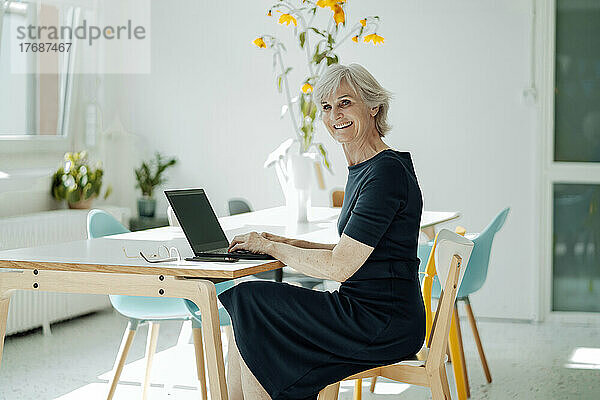 Glückliche Geschäftsfrau mit Laptop sitzt auf einem Stuhl am Schreibtisch im Büro