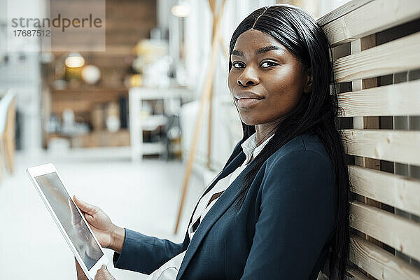 Lächelnde Geschäftsfrau mit Tablet-PC  die sich auf eine hölzerne Heizkörperabdeckung stützt