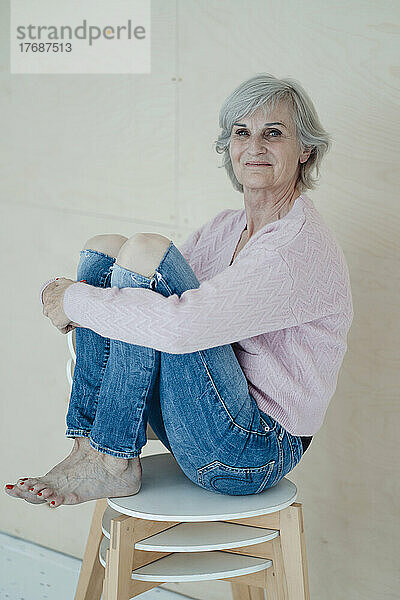Lächelnde ältere Frau mit grauen Haaren sitzt auf Stühlen vor der Wand
