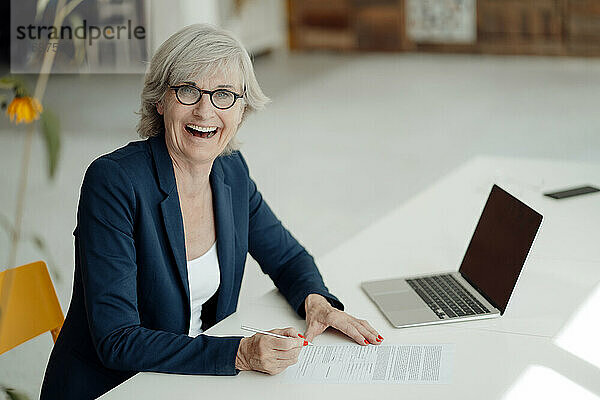 Fröhliche Geschäftsfrau mit Dokument sitzt am Schreibtisch im Büro