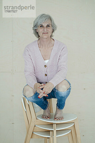 Lächelnde ältere Frau mit grauen Haaren kauert auf Stühlen vor der Wand